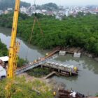 Imagem áerea mostra um guindaste posicionando uma grande viga em estruturas de apoio de uma construção localizada em um rio, com vegetação nas margens