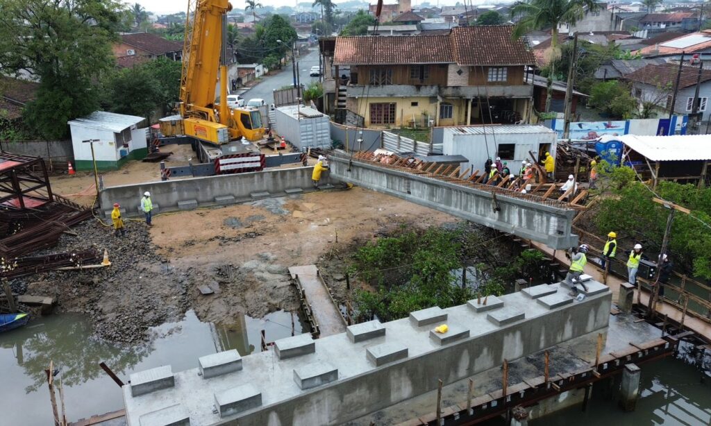 Imagem aérea mostra uma viga sendo posicionada nos apoios de construção de uma ponte. Há pessoas e outras máquinas no entorno