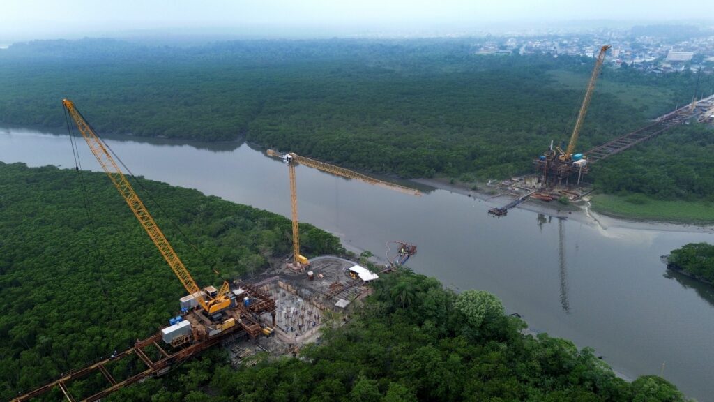 Imagem aérea mostra rio com entorno em vegetação. Há guindastes e equipamentos nas duas margens