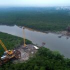 Imagem aérea mostra rio com entorno em vegetação. Há guindastes e equipamentos nas duas margens