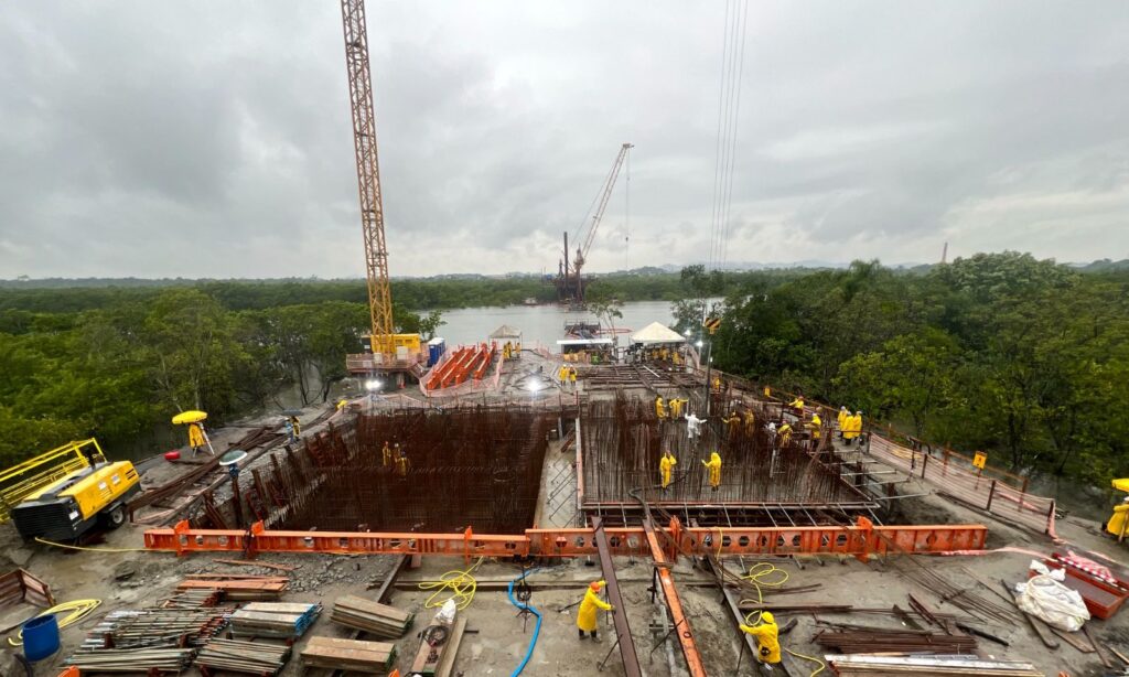 Imagem mostra grande construção, com guindaste, área com armaduras de ferro e outra área com concretagem de espaço com ferro armado. Ao fundo, há um rio, vegetação e outro guindaste
