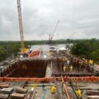 Imagem mostra grande construção, com guindaste, área com armaduras de ferro e outra área com concretagem de espaço com ferro armado. Ao fundo, há um rio, vegetação e outro guindaste