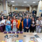 Em um auditório, nove pessoas, entre homens e mulheres, estão posicionadas à frente, seguram troféus e sorriem para a foto. Ao fundo, há dezenas de pessoas sentadas no auditório.