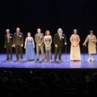 Em um palco iluminado, homens e mulheres idosos e vestidos em traje social posam para o público