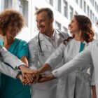 Cinco pessoas, entre homens e mulheres, juntam as mãos. Eles vestem uniformes hospitalares