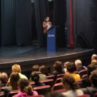 Mulher vestida de calça e jaqueta marrom está em um palco, atrás de um púlpito azul, falando em um microfone. Na plateia, dezenas de homens e mulheres a ouvem