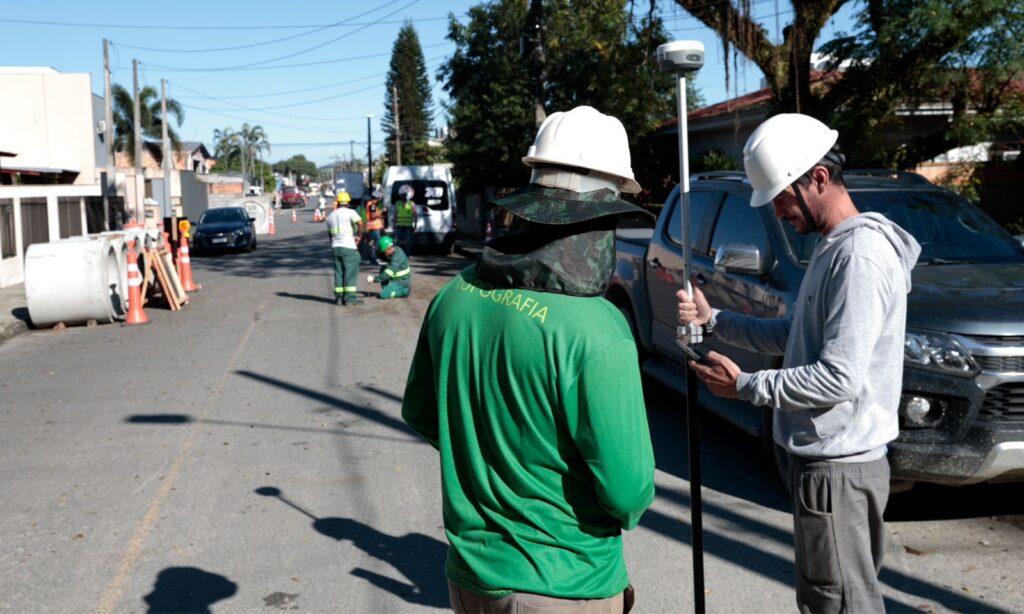 Imagem mostra dois homens com capacetes manuseando equipamentos de topografia. Ao fundo, há outros trabalhadores uniformizados e tubos de concreto em uma rua asfaltada