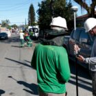 Imagem mostra dois homens com capacetes manuseando equipamentos de topografia. Ao fundo, há outros trabalhadores uniformizados e tubos de concreto em uma rua asfaltada