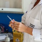 Imagem mostra uma profissional de saúde com jaleco branco. Ela segura uma seringa nas mãos. Ao fundo, há materiais usados para vacinação