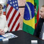 Mulher loira está sentada na ponta de uma mesa. Na lateral, um homem ruivo está sentado. Os dois se olham e conversam. Ao fundo, há bandeiras dos EUA, do Brasil e de Santa Catarina.
