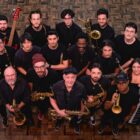 Imagem mostra músicos vestidos de preto com seus instrumentos. Eles estão em pé e olham para a câmera que está num ponto acima