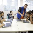 Em uma sala de aula, alunos estão sentados em cadeiras, em carteiras com notebooks. Um professor está em pé e orienta um dos alunos