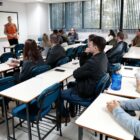 Em um sala de aula, um homem loiro e alto, usando um colete laranja, fala à frente do quadro. Na frente dele, há diversas pessoas sentadas em cadeiras com carteiras escolares.