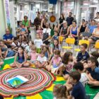 Imagem mostra o espaço de uma biblioteca com dezenas de crianças sentadas e adultos em pé, prestando atenção a um mesmo ponto