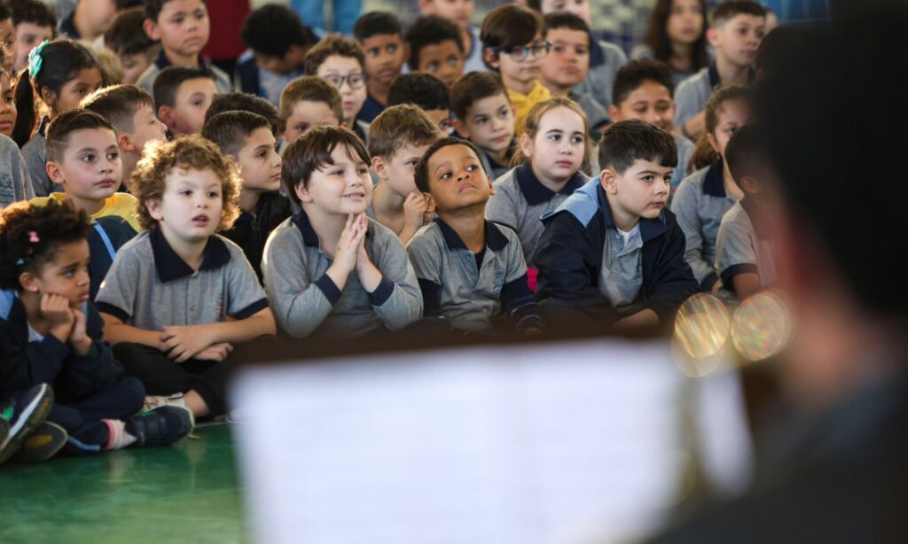 Crianças de cerca de seis anos estão sentadas no chão de um ginásio. Elas parecem concentradas observando uma apresentação de música que ocorre no local