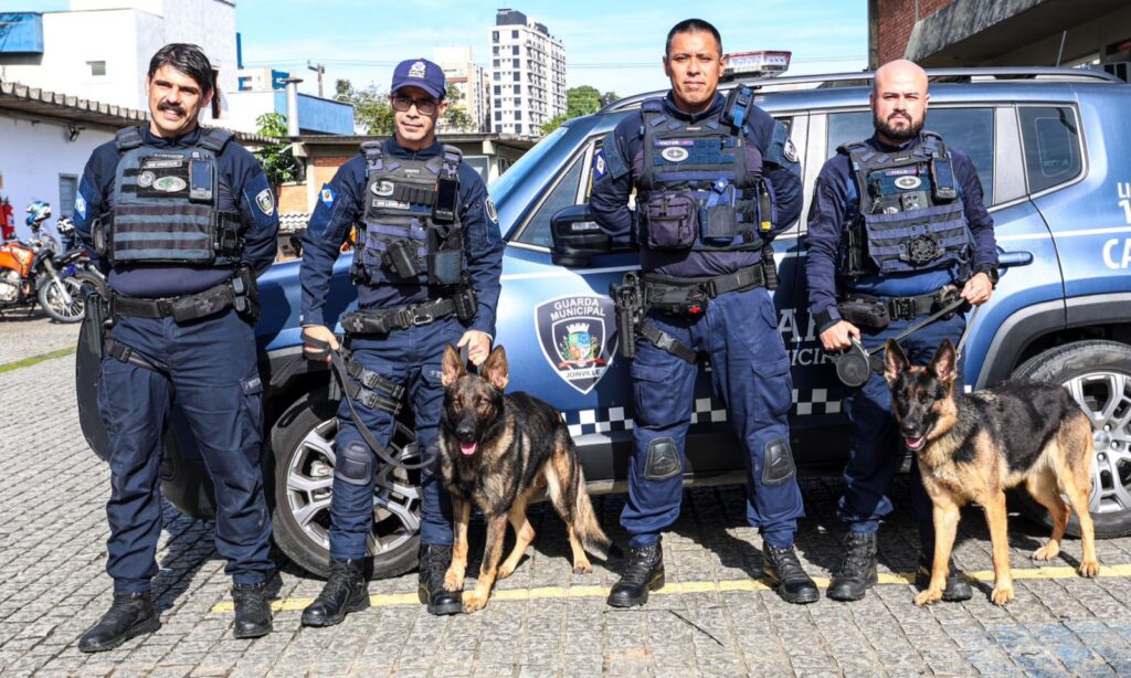 Quatro homens com uniforme da Guarda Municipal posam para foto na frente de um carro da corporação. Dois deles estão segurando a guia de dois cães da raça Pastor Alemão, que estão em pé