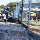 Dois homens uniformizados trabalham na obra de uma calçada perto de um CEI. Eles seguram pás em um terreno com brita e lona, perto de um carrinho de mão