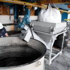 Na imagem, há um grande tanque com líquido preto e um homem com capacete o observando. Perto do tanque, há um equipamento com um grande saco de carvão em cima