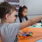 Em primeiro plano, um menino brinca com peças de lego. Ao fundo, outra menina também está brincando