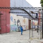 Imagem mostra andaimes ao lado de um grande pavilhão. Há um homem em cima de um deles e outros homens trabalhando no chão
