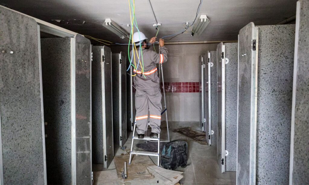 Imagem mostra homem uniformizado em cima de uma escada manuseando a fiação do teto de um banheiro em obras