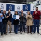 Homens e mulheres estão reunidos em pé, à frente de um painel azul e branco com logo da Prefeitura. Uma mulher loira segura um documento e o exibe para a foto enquanto os demais sorriem e aplaudem