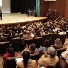 Imagem mostra um auditório cheio com dezenas de pessoas sentadas e duas pessoas no palco