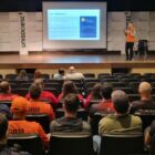 Em um auditório, homem branco está em pé com o microfone no palco, falando ao lado de um telão. Na plateia, há homens e mulheres sentados observado a apresentação de costas para a câmera