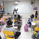 Em sala de aula, há vários alunos sentados em cadeiras, enquanto a professora está à frente da sala, gesticulando
