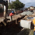 Imagem mostra um homem e uma máquina escavadeira trabalhando próximos a uma grande vala em uma estrada de chão. Há um tubo de concreto e um monte de brita no local