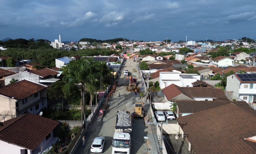 Imagem aérea mostra caminhão de pedra e máquinas em uma estrada de chão com residências