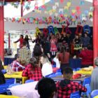 No pátio de uma escola, há decoração de bandeirinhas coloridas. Em um palco, crianças vestidas com roupas típicas de festa junina dançam. À frente do palco, há mesas e pessoas assistindo à apresentação.