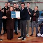 Em uma quadra esportiva, uma mulher e cerca de dez homens sorriem para a foto e aplaudem. No centro, a mulher e um dos homens seguram um documento