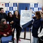 Imagem mostra homens e mulheres em pé e uma mulher em uma cadeira de rodas ao lado de uma placa de metal com o termo Inauguração do CRAS Pirabeiraba. As pessoas sorriem.