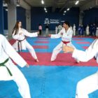 Quatro adolescentes, um menino e três meninas, estão vestidos com uniformes de karatê. Eles estão divididos em duas duplas, frente a frente, em posição de luta