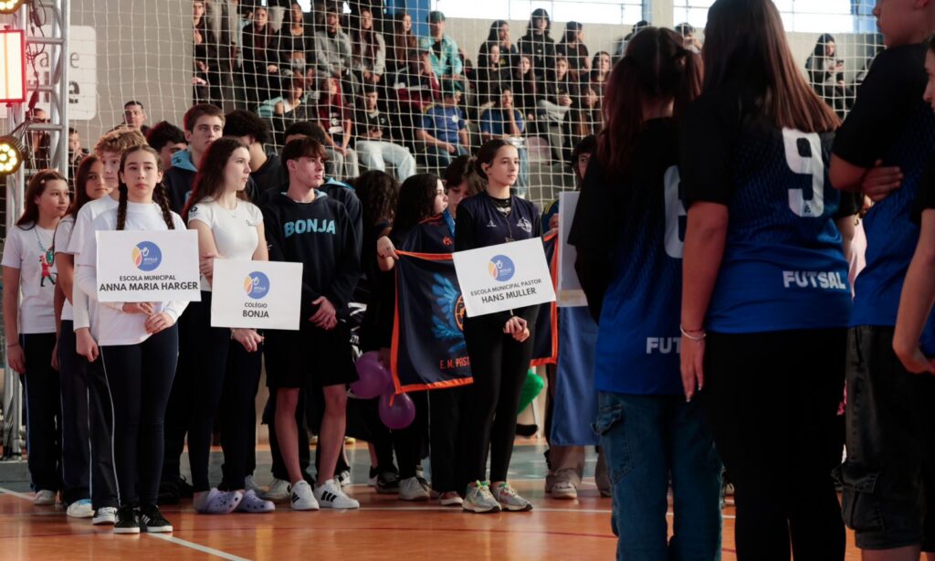 Imagem mostra adolescentes em pé, em uma quadra de esportes. Alguns deles seguram uma placa com o nome de escolas