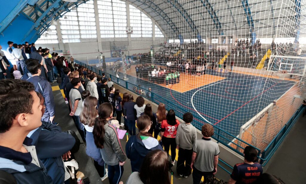 Imagem mostra um ginásio de esportes lotado, com pessoas na quadra e nas arquibancadas