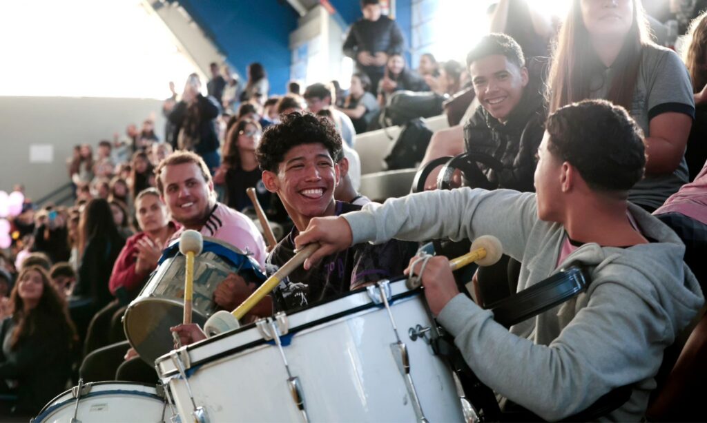 Meninos sorriem enquanto tocam um instrumento de percussão em uma arquibancada com outras pessoas