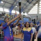Mulher grisalha está em pé e sorri enquanto segura uma tocha. Perto dela, outros idosos observam e sorriem