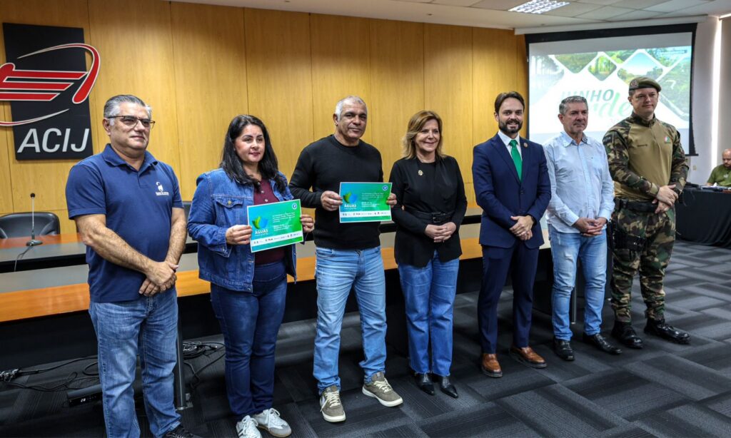 Duas mulheres e cinco homens estão em pé, lado a lado, e posam para foto. Ao centro, um homem e uma mulher seguram um documento