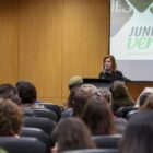 Uma mulher loira está à frente de uma sala, onde fala em um púlpito. Atrás dela, há um painel escrito junho verde