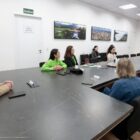 Imagem mostra grande mesa de reuniões ocupada por algumas pessoas, a maioria mulheres, assistindo a uma apresentação na televisão à frente da sala