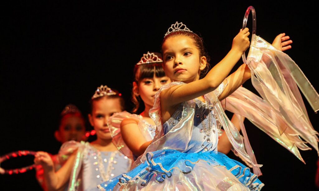 Meninas com figurino de ballet estão em pé, em um palco. Elas usam tiaras na cabeça e têm expressão séria