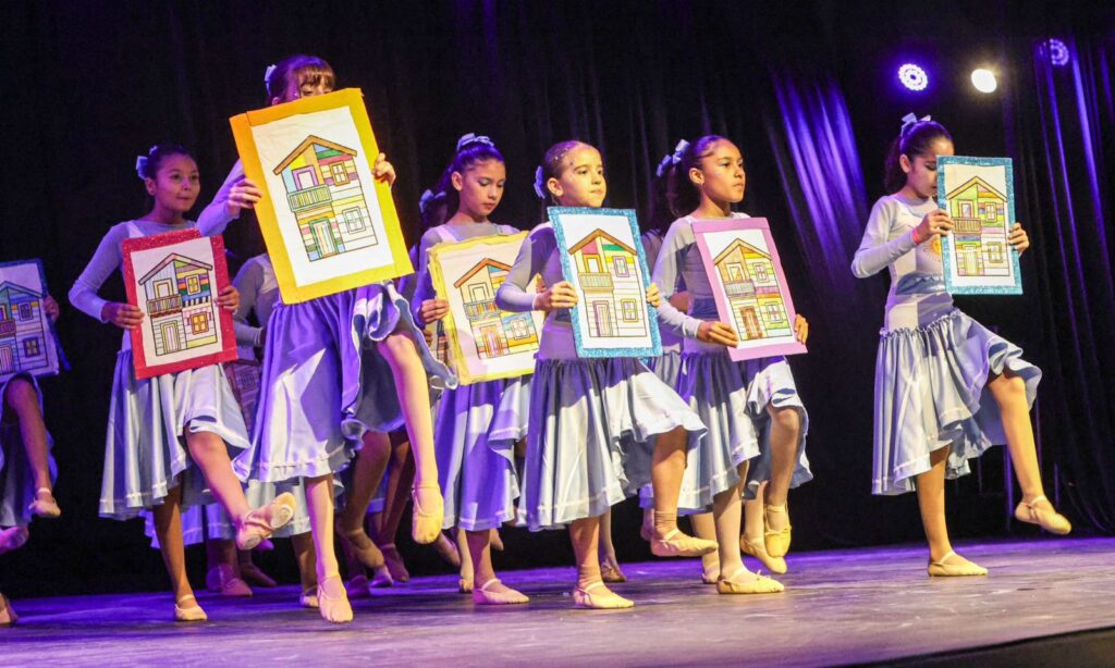 Crianças com figurino de dança se apresentam em um palco. Elas seguram cartazes com desenhos de casa