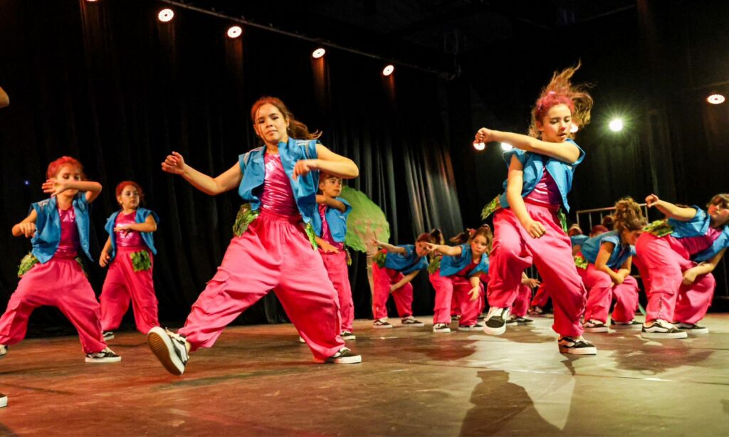 Grupo de adolescentes com figurino rosa, verde e azul dança num palco
