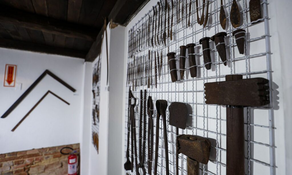 Imagem mostra tela aramada instalada em uma parede branca onde estão penduradas várias ferramentas manuais para fazer esculturas
