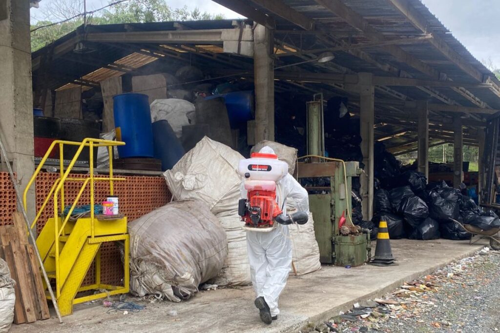 Homem com macacão de proteção branco carrega um pulverizador nas costas. Ele está caminhando perto de um galpão onde há vários sacos de lixo.