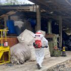 Homem com macacão de proteção branco carrega um pulverizador nas costas. Ele está caminhando perto de um galpão onde há vários sacos de lixo.