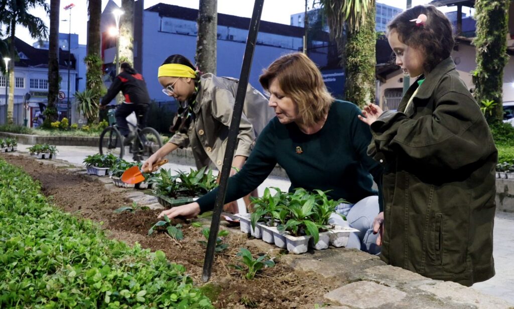 Mulher loira de blusa verde manuseia planta em um canteiro. Ao lado dela, há duas meninas crianças e, ao fundo, um homem passeando de bicicleta
