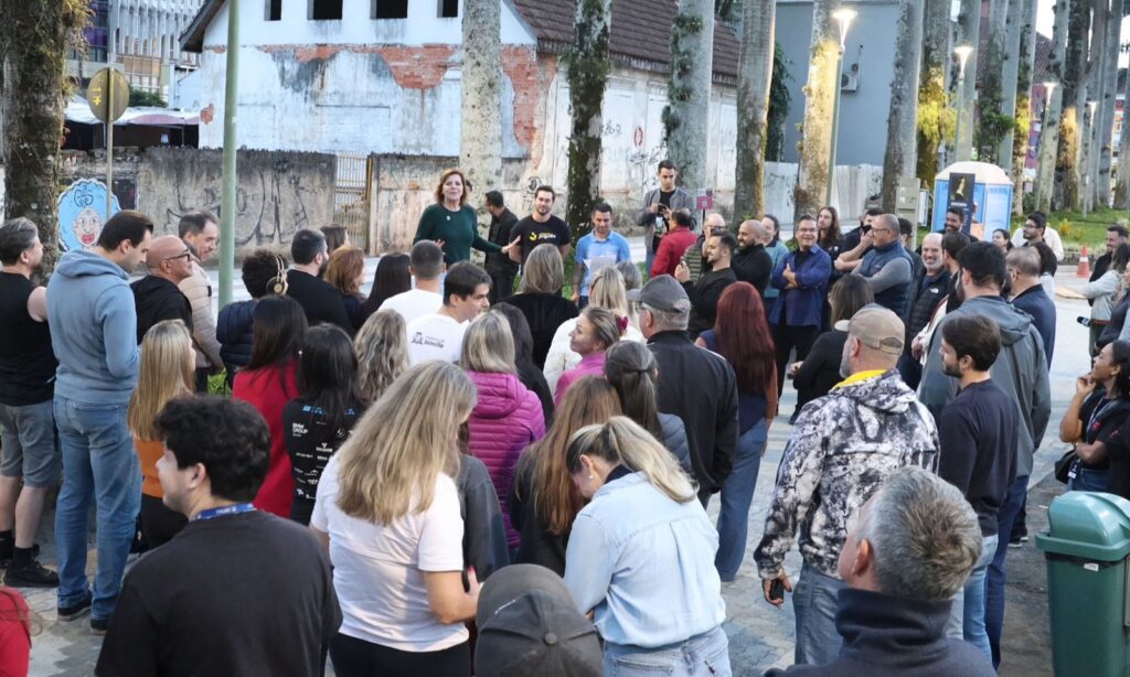 Mulher loira de camisa verde fala e gesticula em meio a dezenas de pessoas que estão ao seu redor em uma rua com árvores